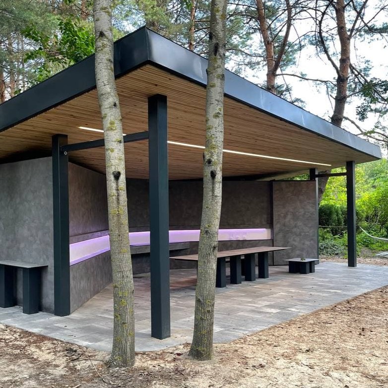 Modern outdoor pavilion with steel-framed roof, wood slat ceiling, and built-in bench seating around existing trees
