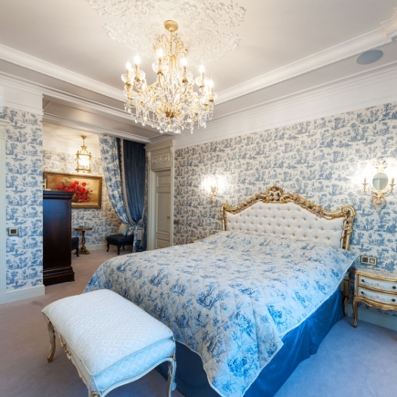 Bedroom with blue toile wallpaper, a gilded tufted headboard, and crystal chandelier