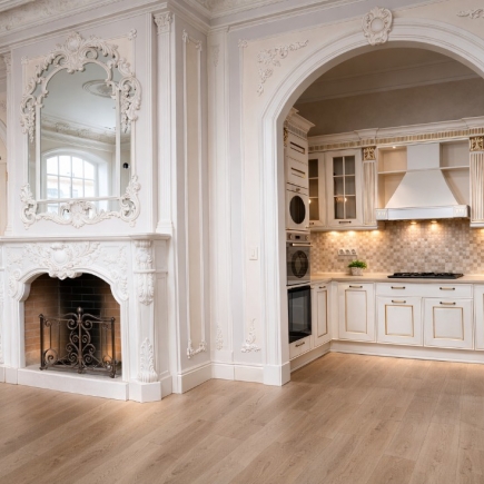 Carved white marble fireplace and mirror beside an arched kitchen doorway with oak flooring