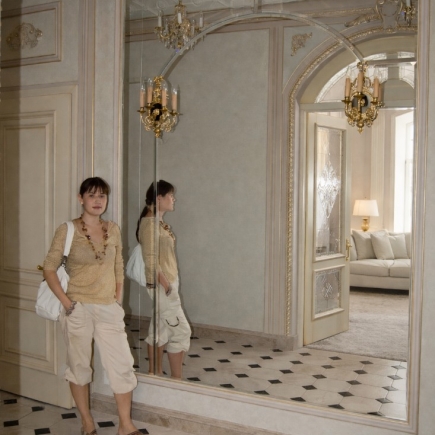 Woman reflected in the mirrored corridor beside gilt wall sconces and an arched doorway