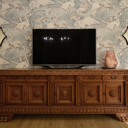 Carved antique sideboard with wall-mounted TV against flamingo-print wallpaper