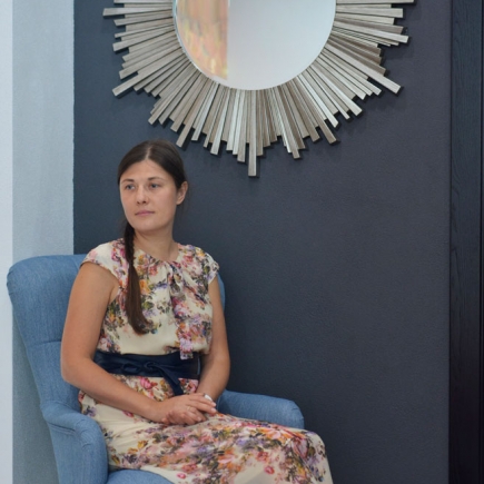 Woman in a floral dress seated in a blue armchair beneath a sunburst mirror on a dark accent wall