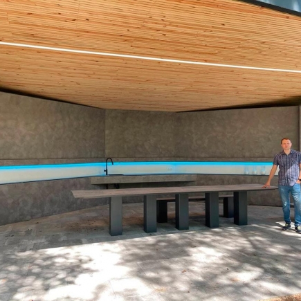 Man standing beside the built-in concrete dining table under the pavilion's wood-slat roof, with a blue LED light strip along the back wall