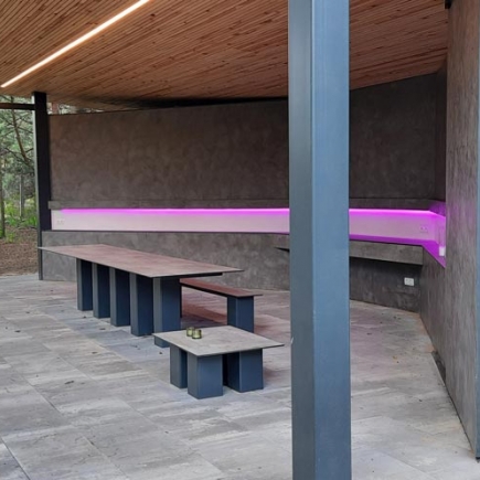 Pavilion interior with built-in table and bench beneath the wood-slat roof, lit by a pink LED strip