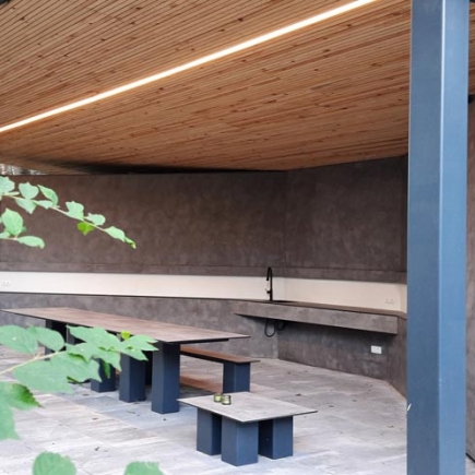 Pavilion interior with built-in table and bench, framed by climbing greenery and a support column