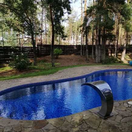 Curved swimming pool with a stainless steel water feature, framed by pines