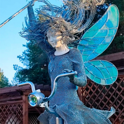 Fairy-on-bicycle sculpture overlooking the white brick cottage and lawn under a cloudy sky