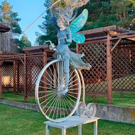Wrought bronze fairy sculpture riding a penny-farthing bicycle in the garden at dusk